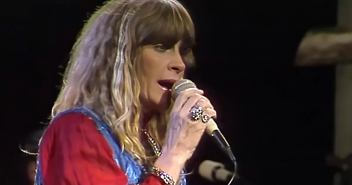 A person with long, wavy hair sings into a microphone, wearing a red and blue outfit.