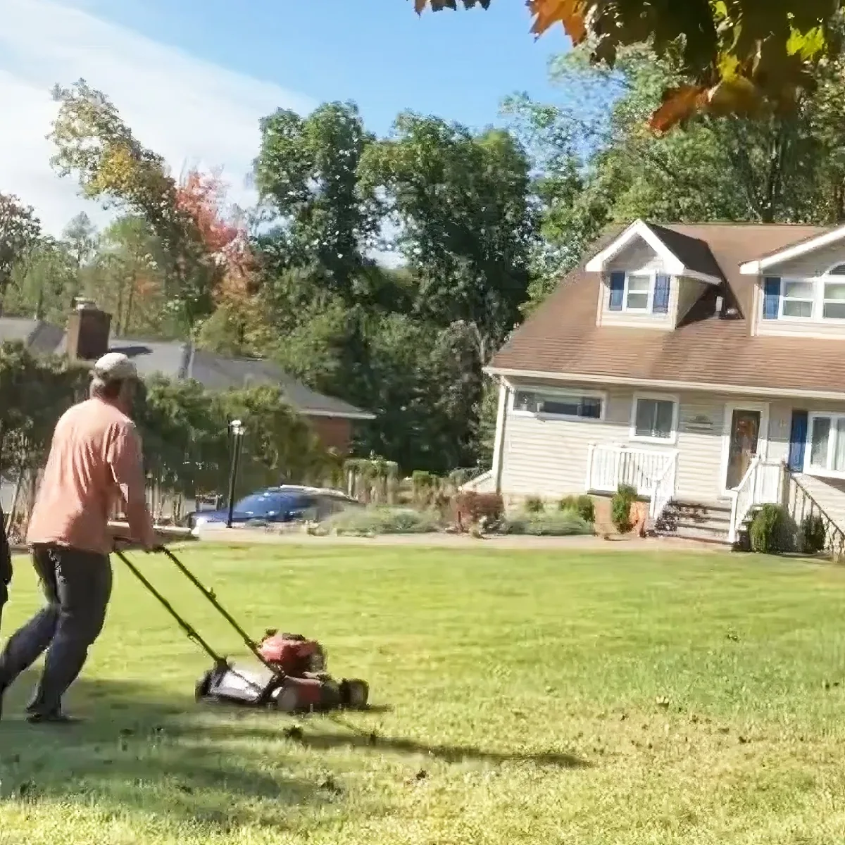 A man mows a lawn in front of a house with a red and black lawnmower.