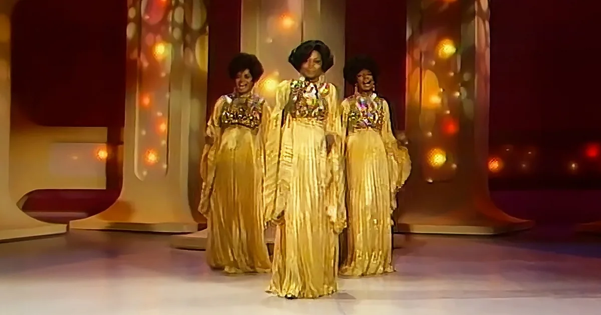 Three women in gold dresses with afros and floral necklaces perform on a stage with colorful lights.