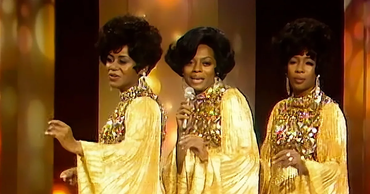 Three women in gold sequined gowns with afro hairstyles perform on stage.