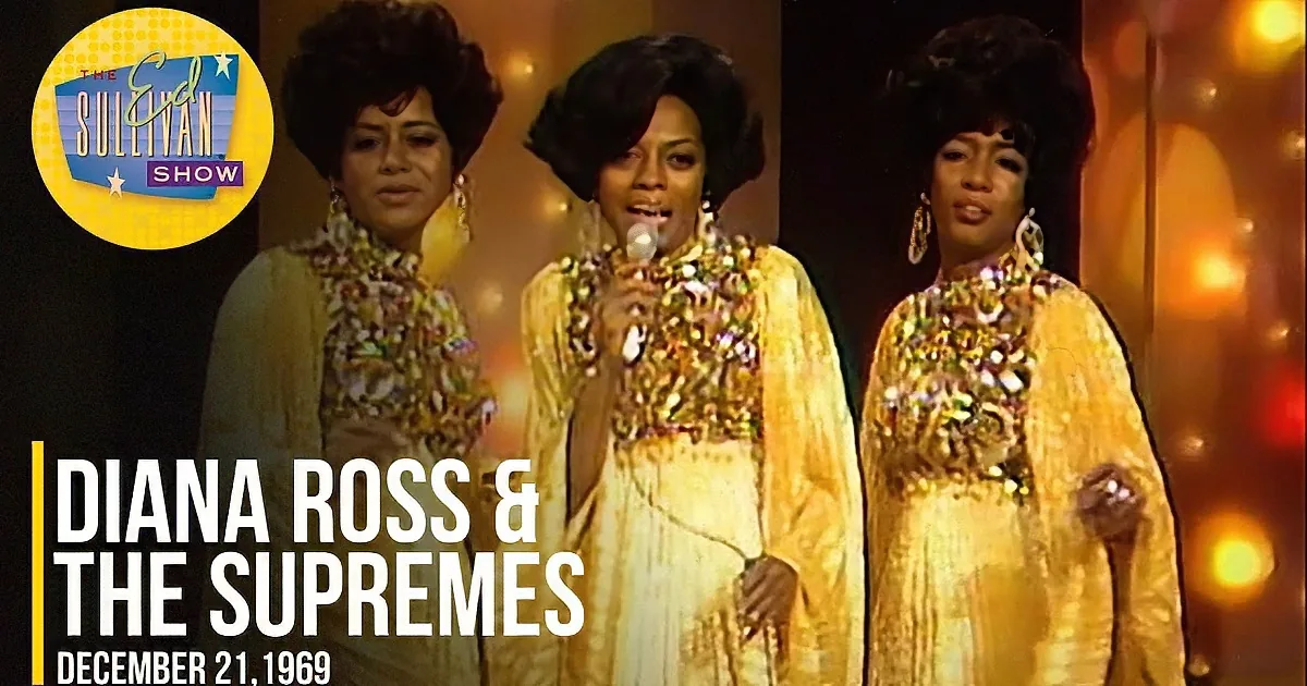 Three women in sequined outfits, smiling, one holding a microphone, on stage.