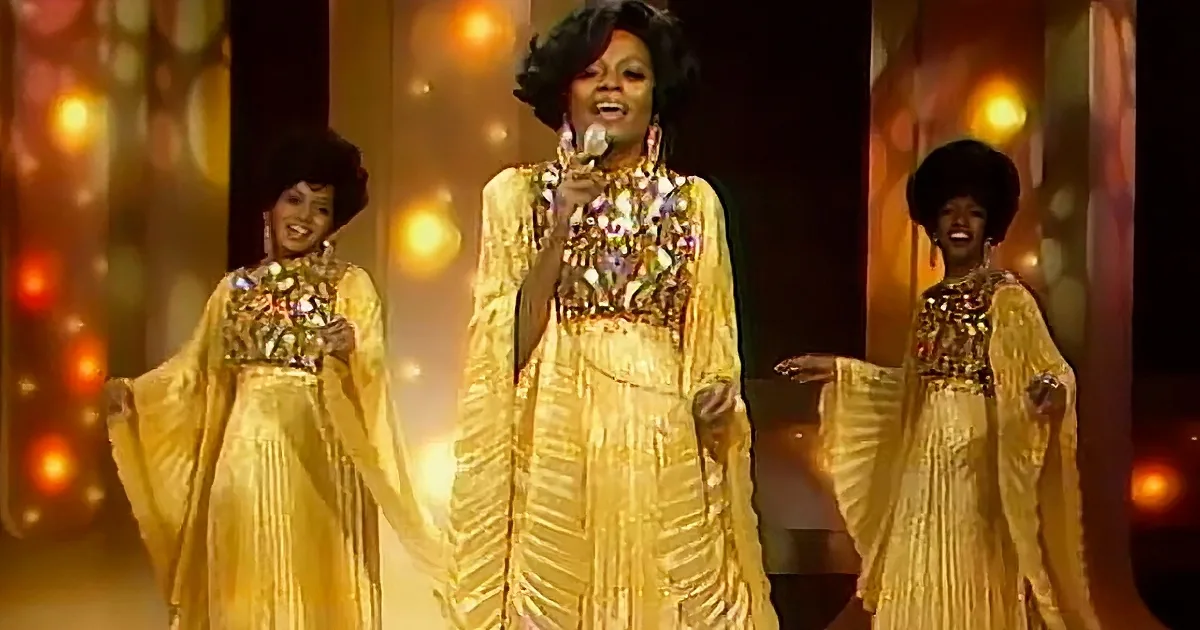 A woman in a yellow floral dress sings on stage with two backup dancers in matching outfits.