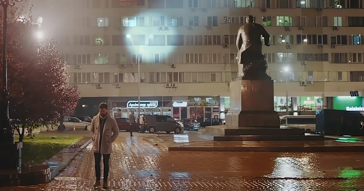 A man in a coat walks on a wet pavement near a statue and illuminated building at night.