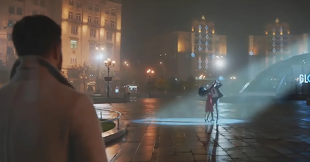 A man walks towards a couple embracing under a fountain in a foggy, illuminated city square.
