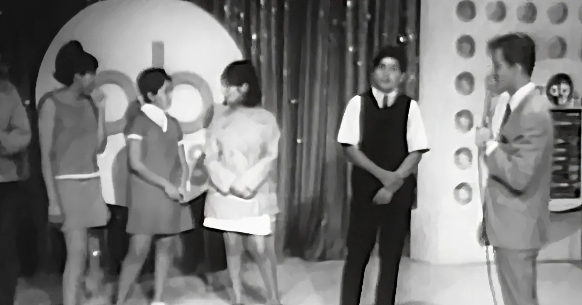 A man in a vest stands with folded arms, surrounded by children in a staged setting with a curtain backdrop.