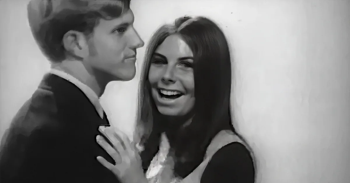 A man and woman are embracing, the woman smiling, both in formal attire, against a plain background.
