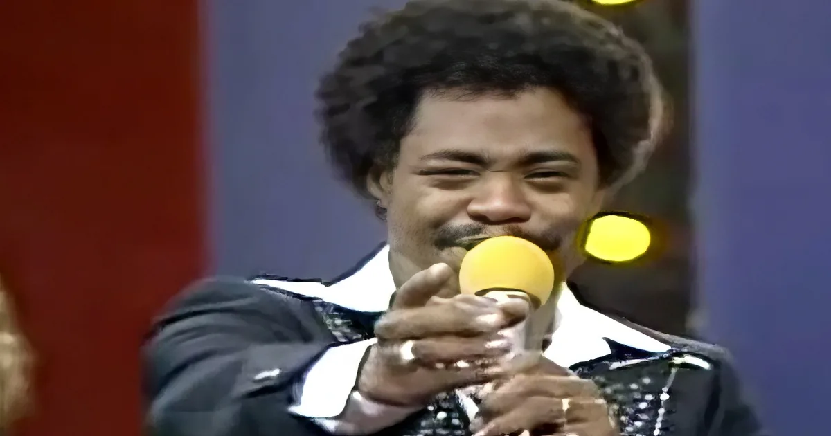 A man in a suit holds a microphone, smiling, with a red and blue background.