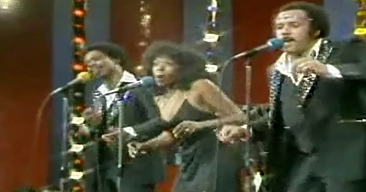 A man in a suit sings into a microphone, accompanied by two other musicians playing instruments.