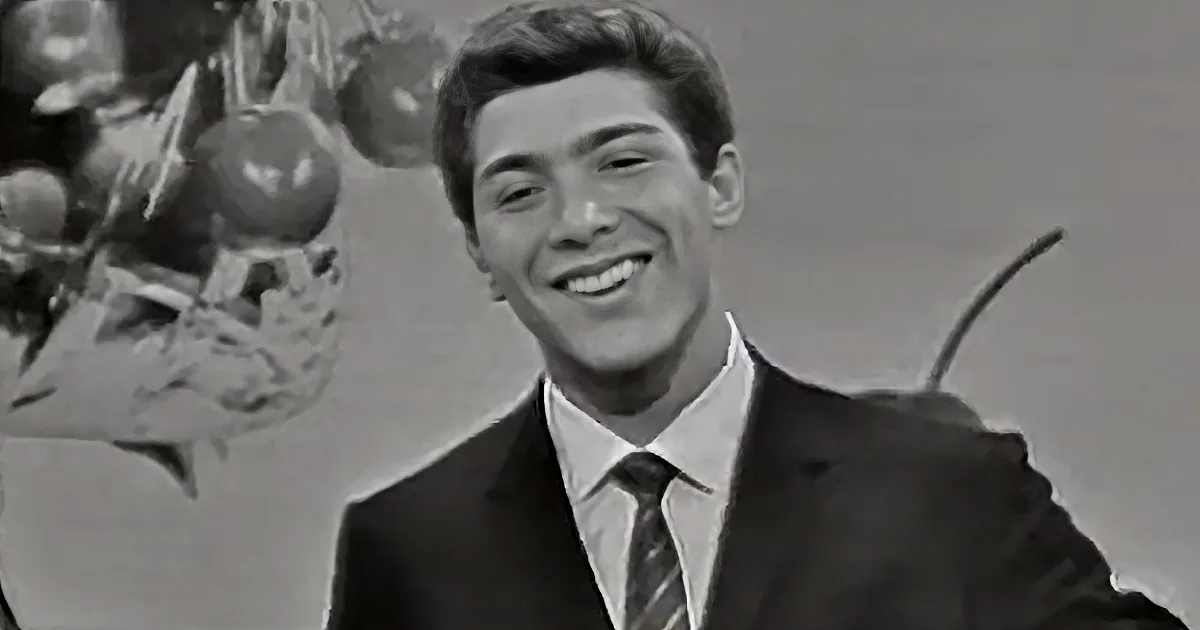 A man in a suit smiles, with a fruit basket and a stick in the background.