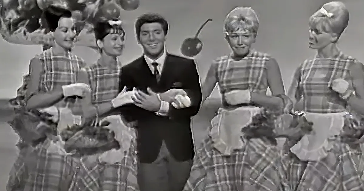 A man in a suit stands with four women in plaid dresses, holding a small object, in a vintage setting.