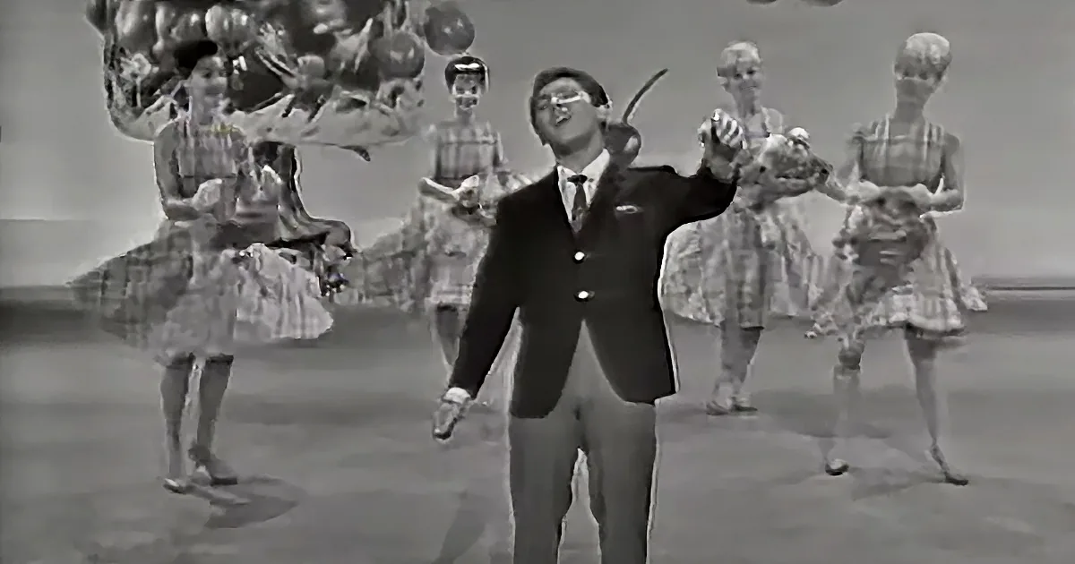 A man in a suit and tie stands center stage, gesturing, with dancers in plaid dresses behind him.