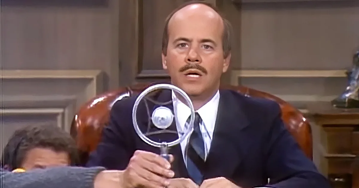 A man in a suit holds a magnifying glass, seated in a leather chair, with a wooden desk and shelves in the background.