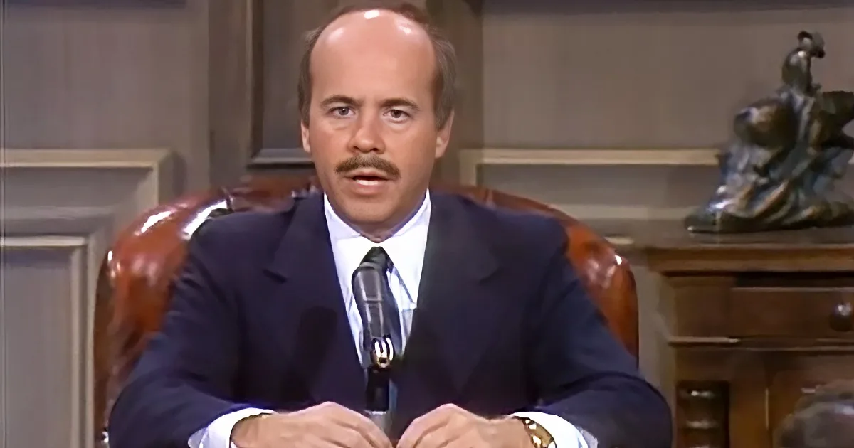 A man in a suit sits at a desk with a microphone, wearing a gold watch, in a formal setting.