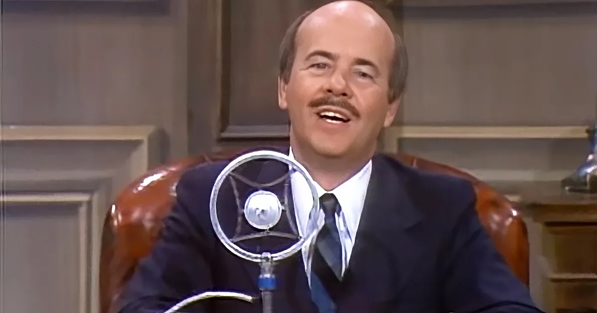 A man in a suit and tie is seated, smiling, with a microphone in front of him.