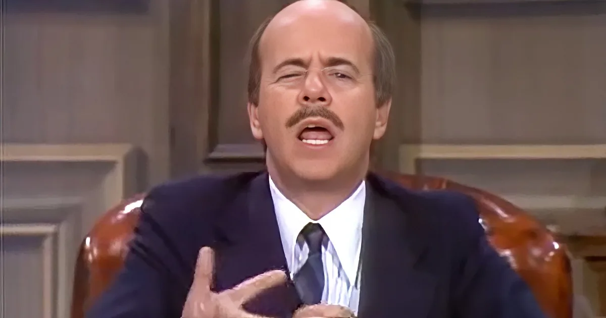A man in a suit and tie is seated, gesturing with his hands while speaking.