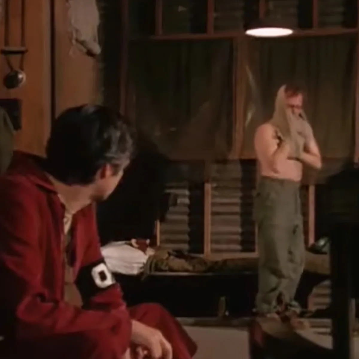 A person in a red outfit sits on a bench, while another stands nearby, covering their face.