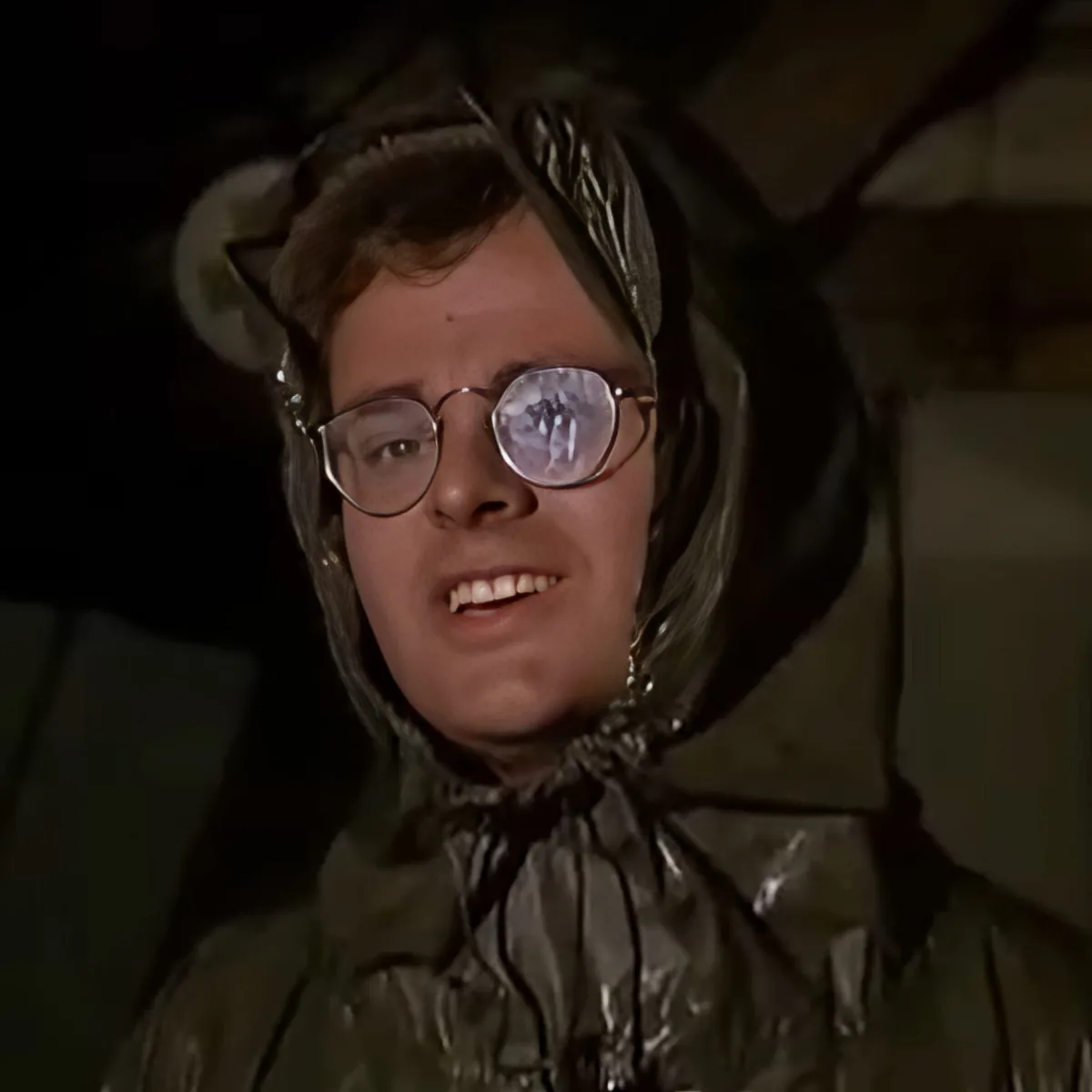 A person wearing a shiny green raincoat and round glasses, smiling, with a dark background.
