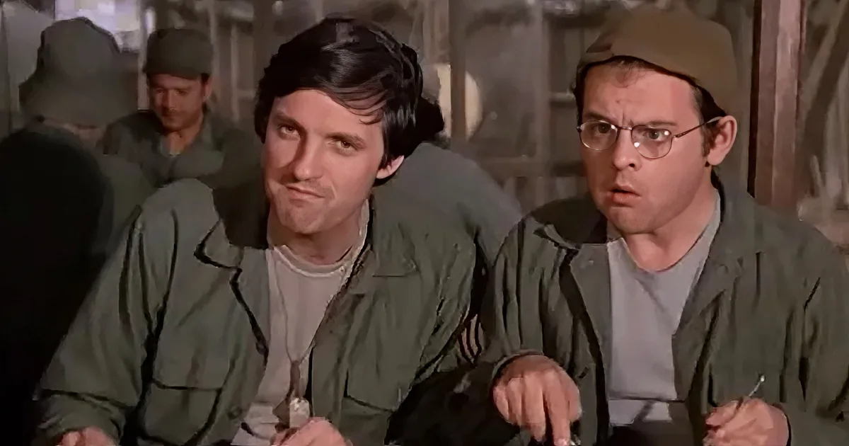 Two men in military uniforms sit side by side, one with a serious expression, the other looking surprised.