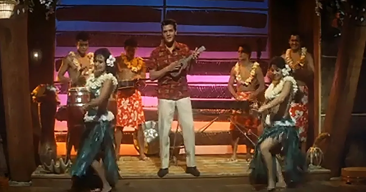 A man in a red shirt plays ukulele on stage surrounded by dancers in colorful attire.