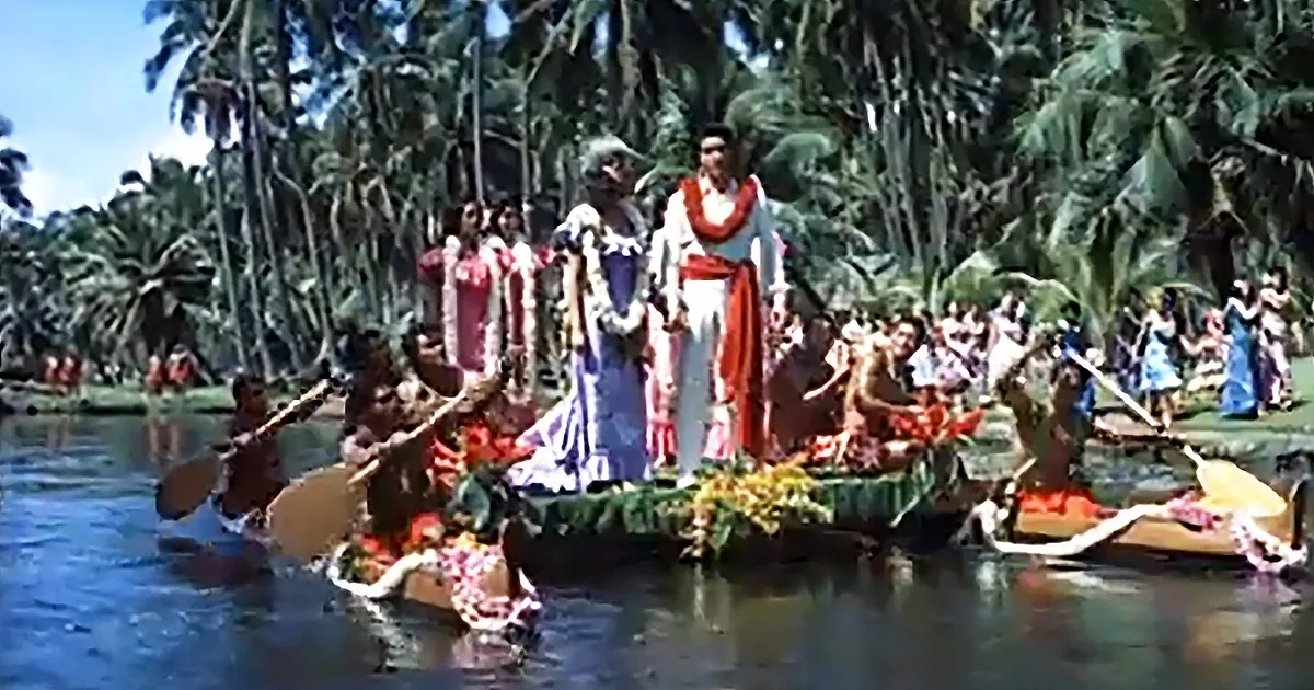 A man in white and red attire stands on a decorated boat surrounded by people in traditional attire and palm trees.