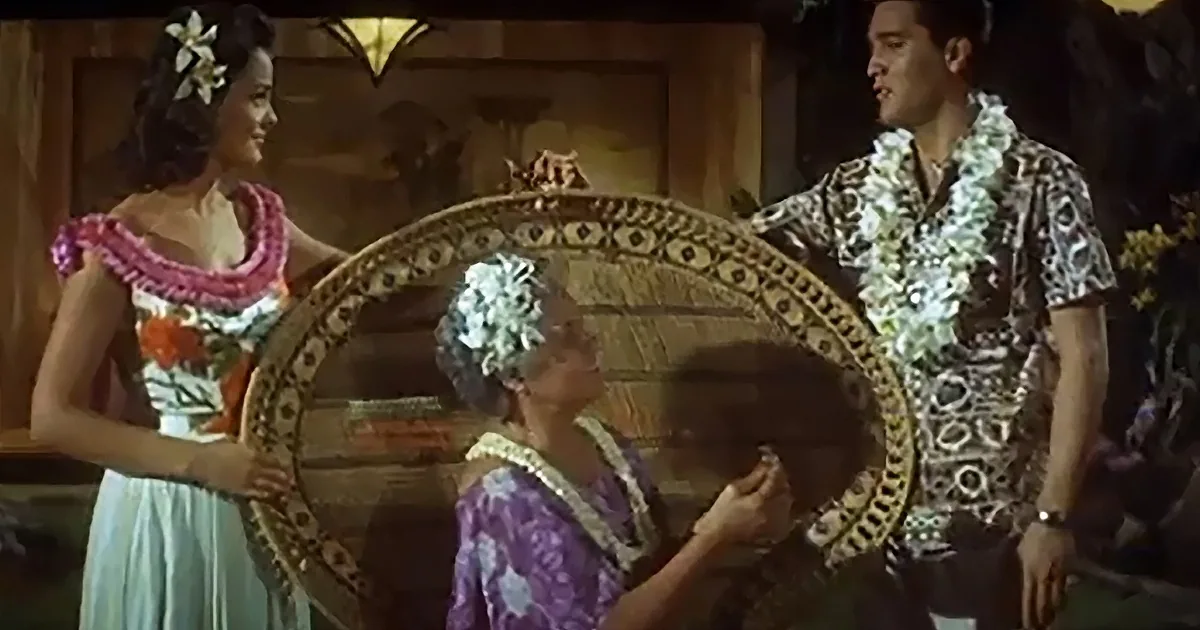 A man and woman in Hawaiian attire hold a large, ornate mirror, surrounded by floral decorations.