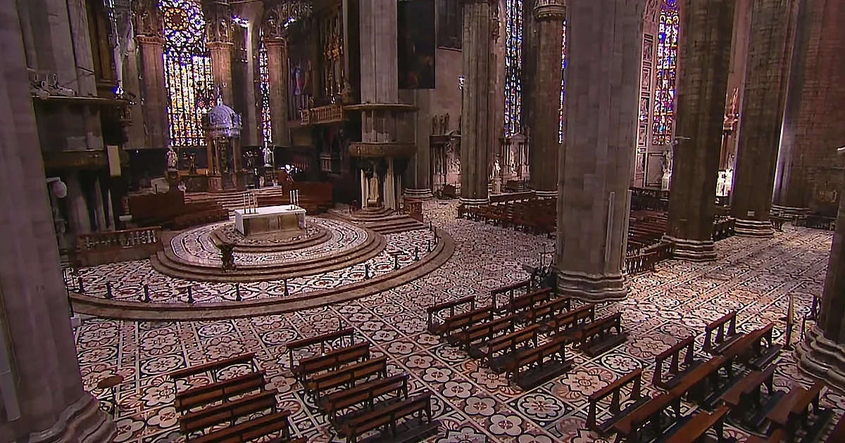 The interior of a grand cathedral with ornate stained glass, intricate floor patterns, and rows of wooden pews.