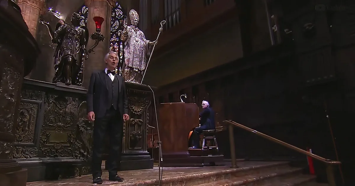 A man in a suit sings on stage with a conductor and statues in the background.