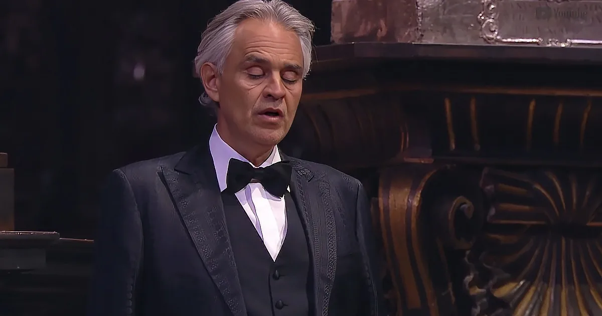 A man in a tuxedo sings with closed eyes, standing in front of ornate furniture.