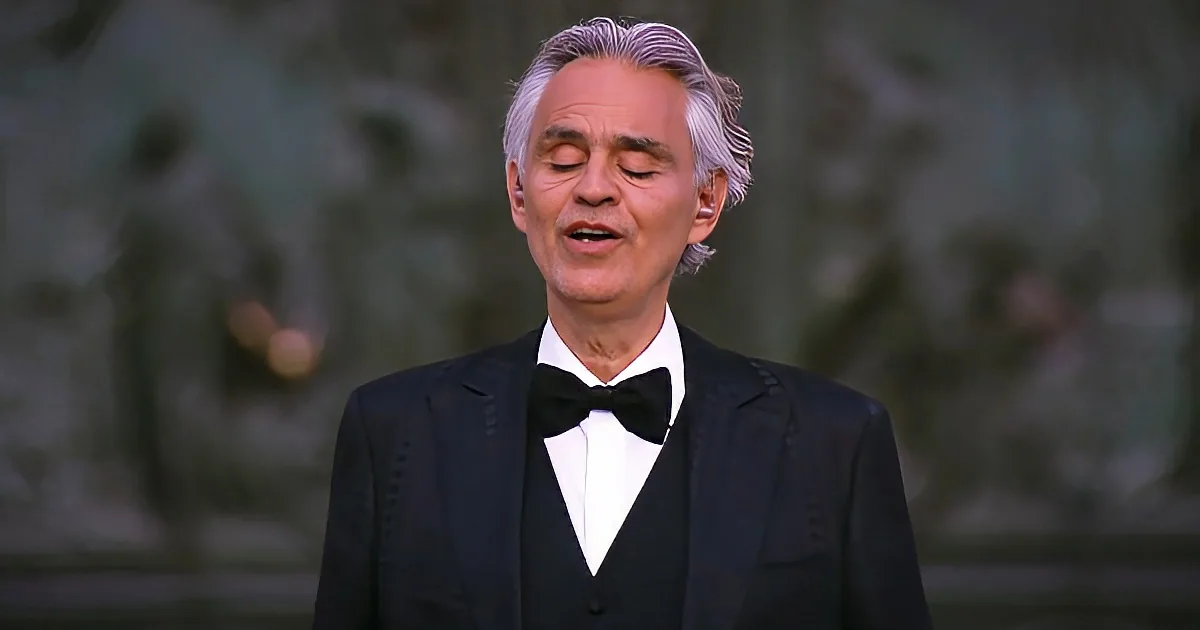 A man in a tuxedo with a bow tie stands with eyes closed, mouth slightly open, in a blurred outdoor setting.