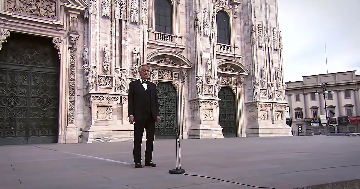A man in a tuxedo stands in front of a grand cathedral with intricate carvings and a microphone nearby.
