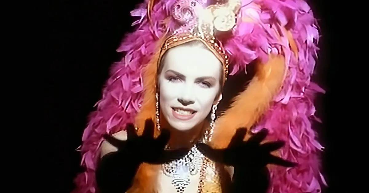 A person in a vibrant, ornate costume with pink feathers and a crown, striking a dramatic pose.