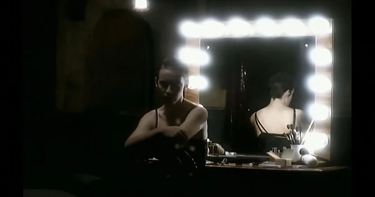 A woman sits in front of a mirror with bright lights, applying makeup in a dimly lit room.