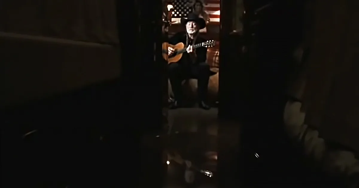 A man in a cowboy hat plays guitar in a dimly lit room with an American flag in the background.