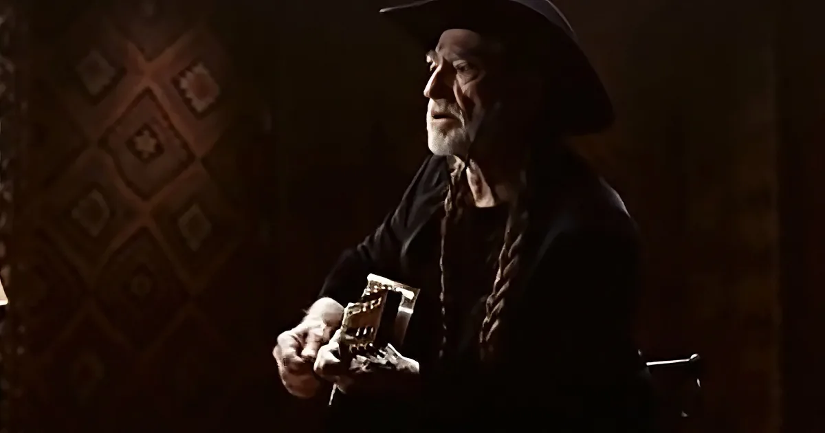 An older man in a cowboy hat holds a guitar, wearing a dark shirt and tie, in a dimly lit room with patterned wallpaper.