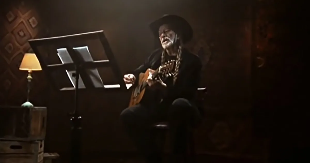 An older man in a cowboy hat plays guitar, surrounded by rustic decor and a lamp.