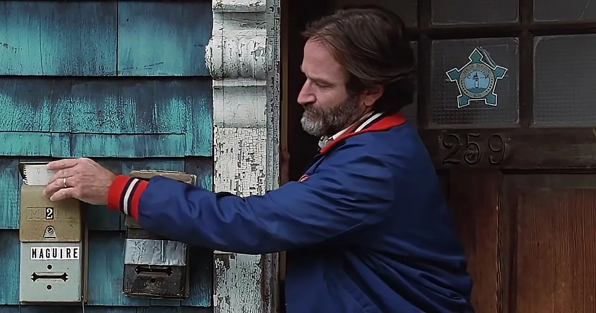 A man in a blue jacket reaches for a mailbox labeled "Maguire" on a weathered house.