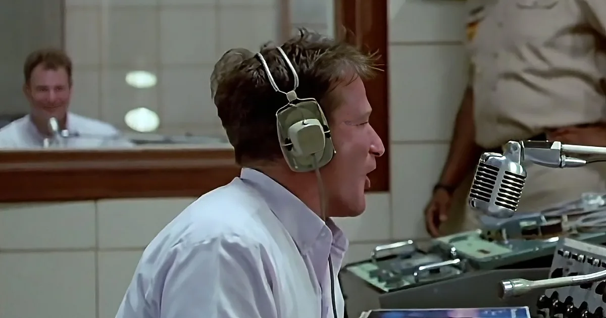 A man wearing headphones and a white shirt is speaking into a microphone in a control room.