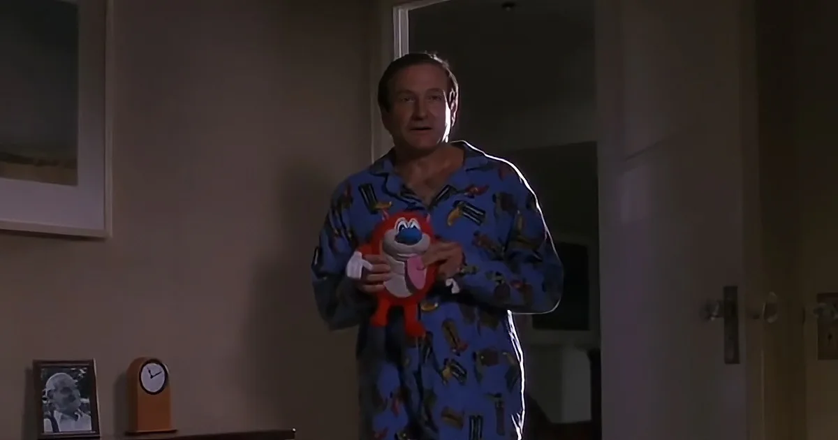 Robin Williams in pajamas holds a toy, standing in a room with a clock and framed picture.