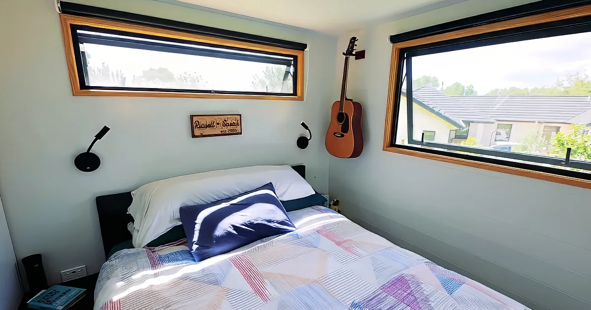 A cozy bedroom with a bed, guitar, and two windows showing a sunny day and a house outside.