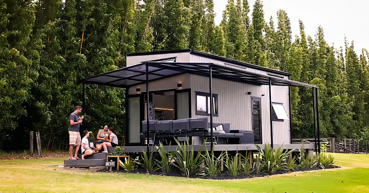 A modern, elevated cabin with a black awning and outdoor seating area surrounded by lush greenery.
