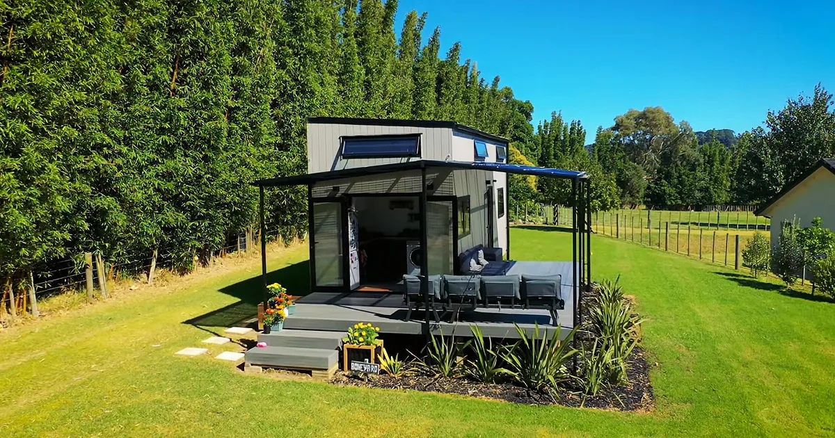 A modern, open-air tiny house with a patio, surrounded by lush greenery and a clear blue sky.