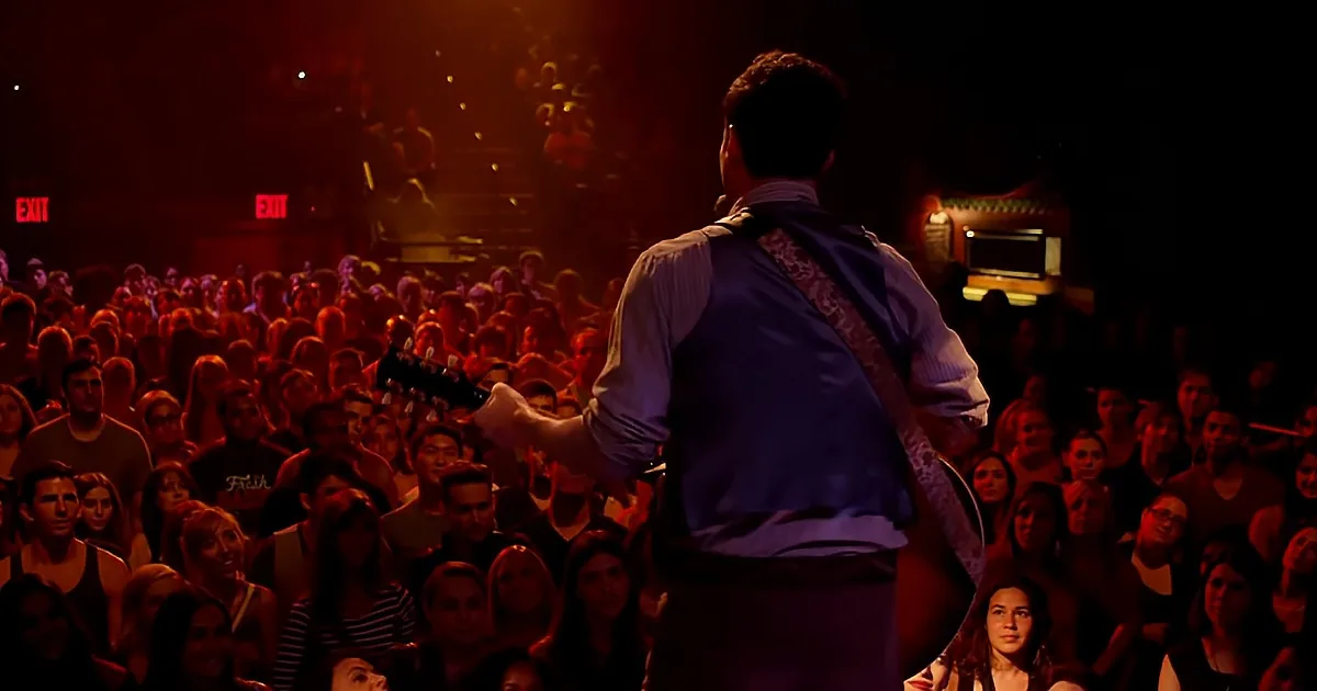 A guitarist in a purple shirt plays on stage in front of a large, engaged audience.