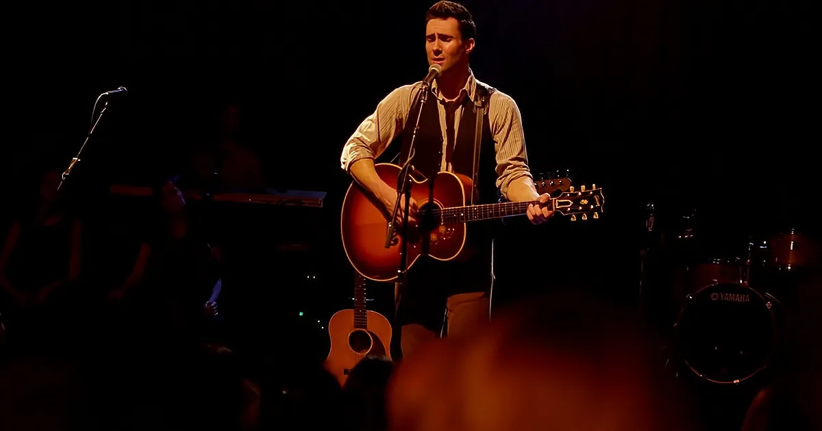 A man plays an acoustic guitar on stage, singing into a microphone, with a band in the background.