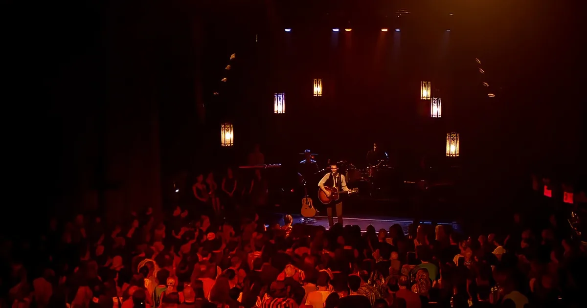 A musician plays guitar on stage under warm lighting, surrounded by a crowd of spectators.