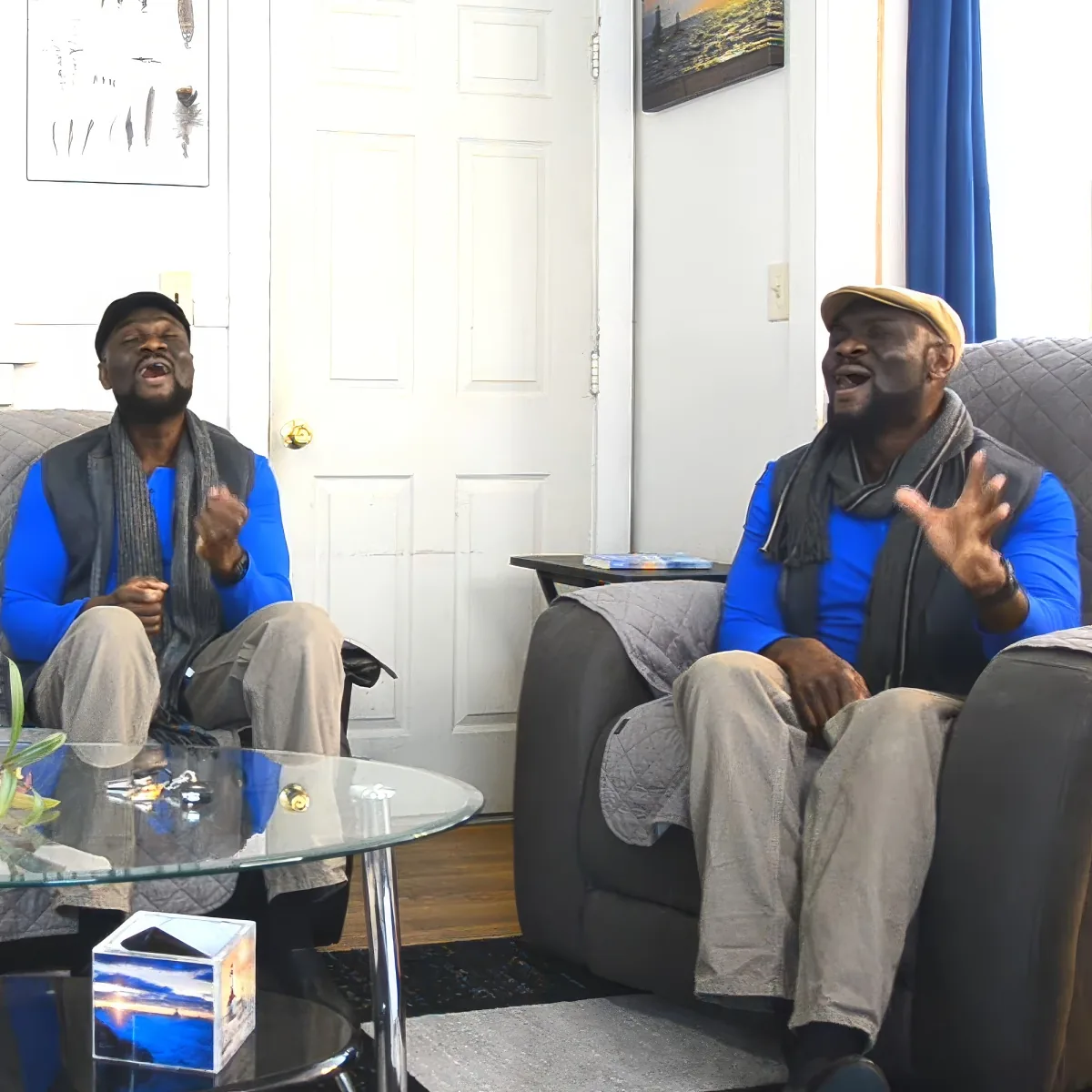 Two men in blue shirts and hats sit on a couch, gesturing and speaking in a living room setting.
