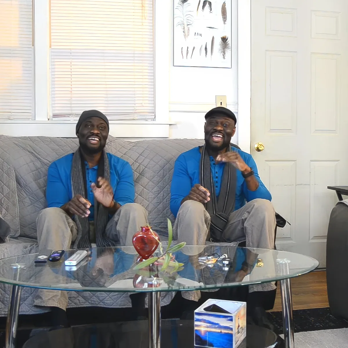 Two men in blue shirts and hats sit on a couch, smiling and gesturing in a living room with a glass table and artwork.