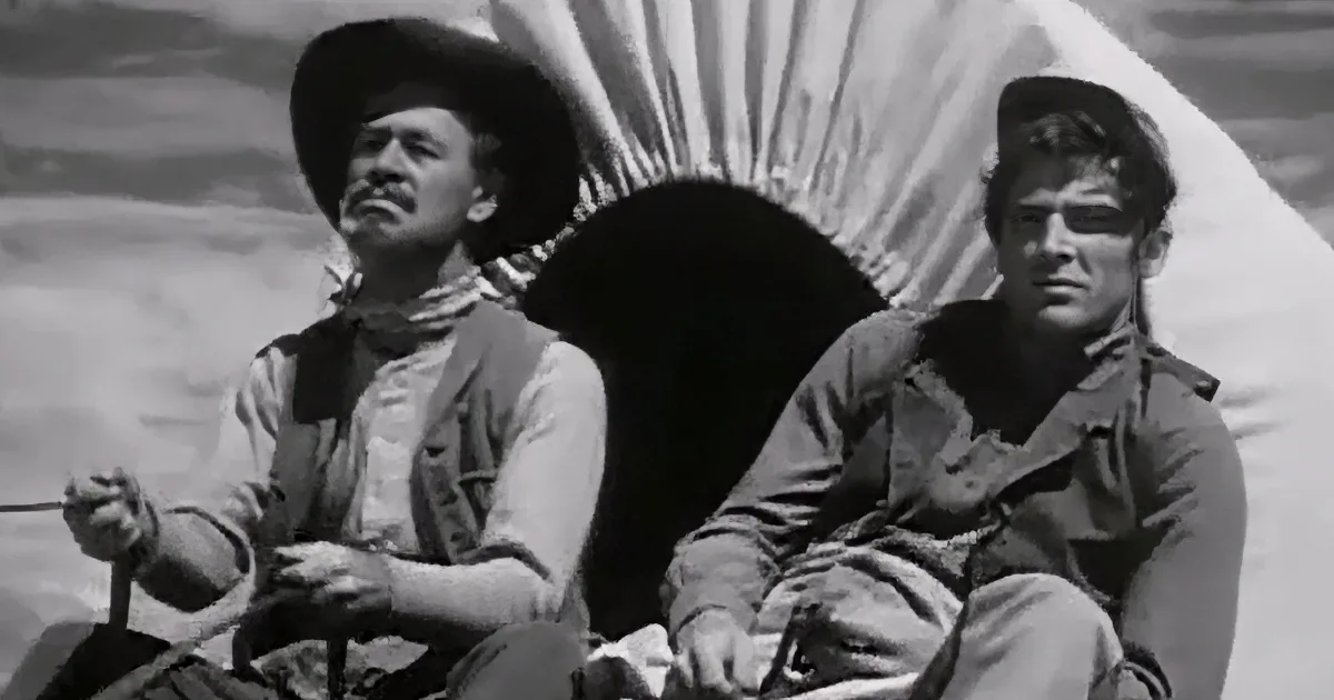 Two men in cowboy attire sit side by side, one holding reins, against a backdrop of a tent and rugged terrain.