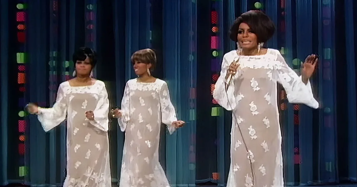Three women in white dresses perform on stage with colorful curtains in the background.