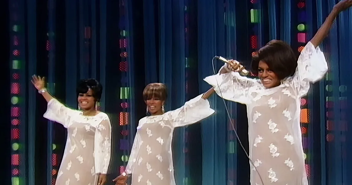 A woman in a white lace dress holds a microphone, smiling, with two others in similar outfits behind her.