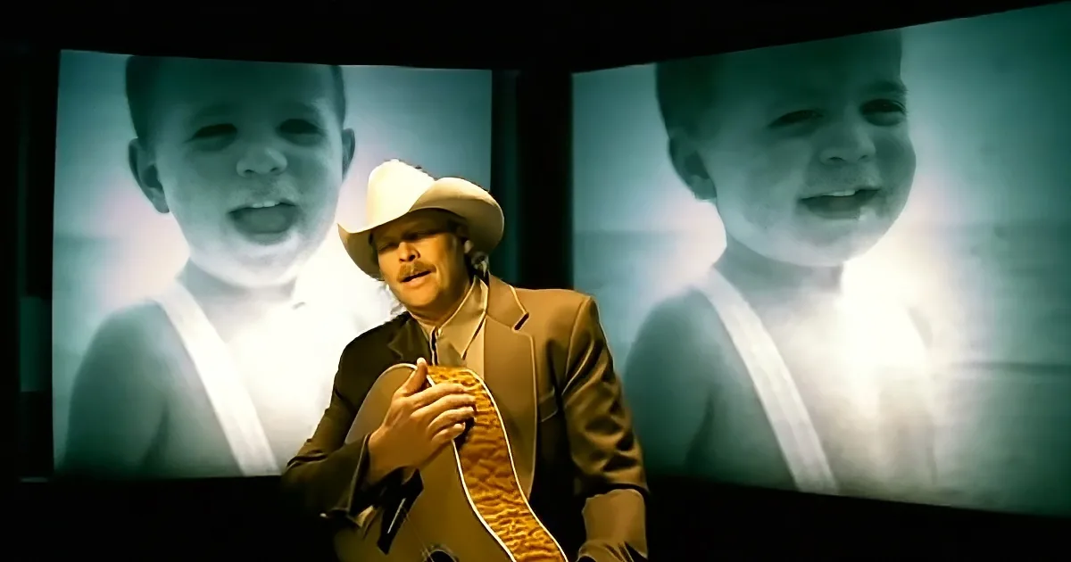 A man in a cowboy hat holds a guitar, with two large screens behind him showing a smiling child.